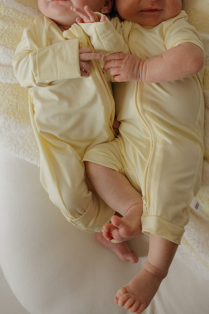 Two babies in matching forever french baby Shortie Bamboo Zip | Sunny rompers lie side by side on a soft white blanket, their arms and legs gently intertwined. The hypoallergenic bamboo fabric ensures their delicate skin stays comfortable and irritation-free.