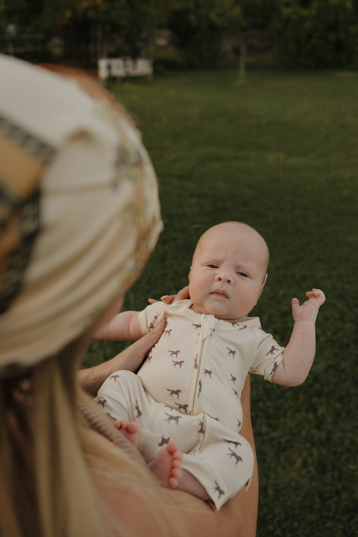 A baby wears the forever french baby Shortie Bamboo Zip in Wild Horses print while being held outdoors by an adult in a patterned headscarf, with grass and trees in the background.