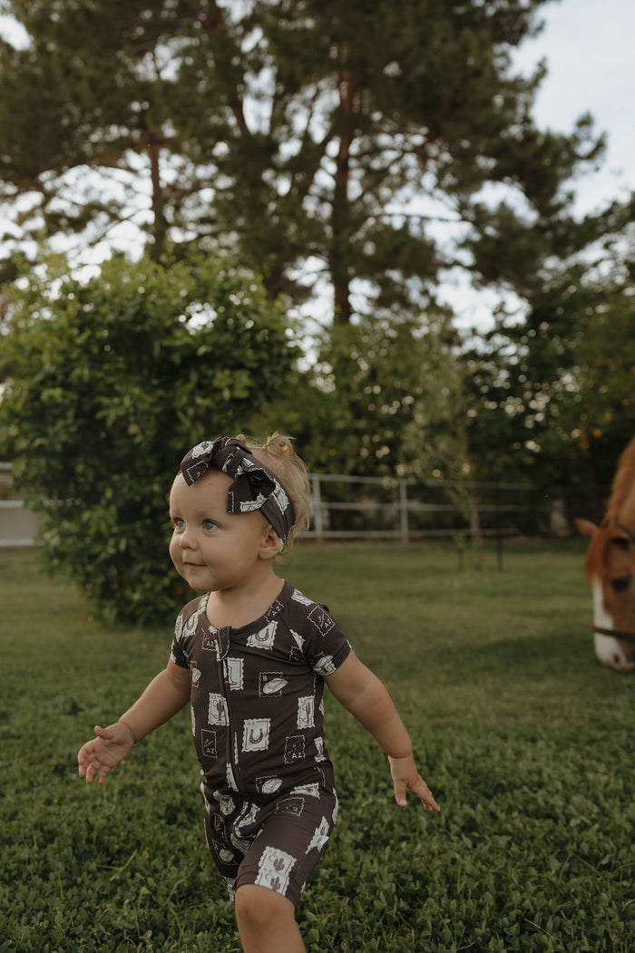 A toddler smiles while walking on grass in a yard, wearing the forever french baby Shortie Bamboo Zip | With Love from AZ pajamas, with a matching headband, as trees and a horse grazing appear in the background.