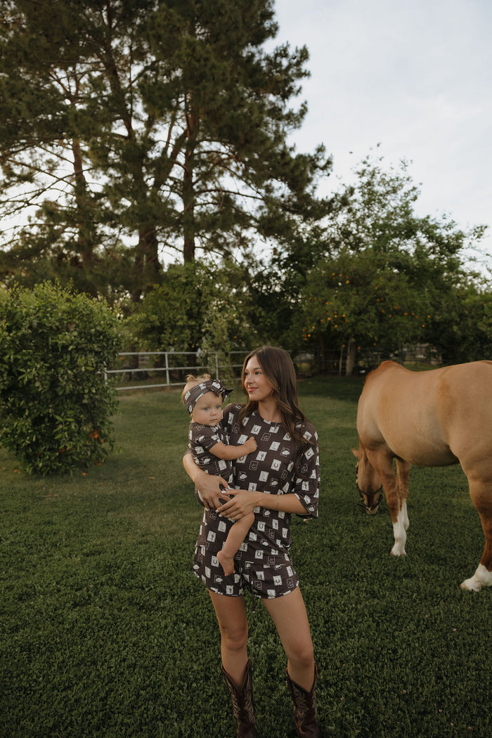 A woman holding a baby stands on grass near a horse. Both wear matching forever french baby Shortie Bamboo Zip pajamas, featuring black and white square patterns. Trees and greenery fill the background under a partly cloudy sky.