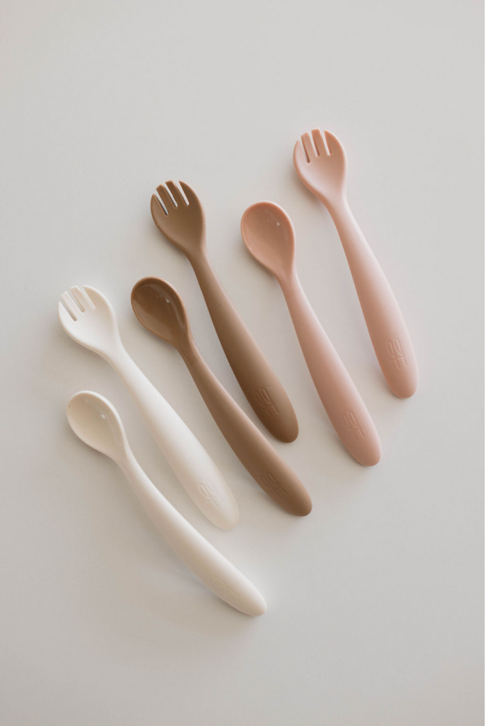 A set of six BPA-free feeding utensils from the Silicone Utensil Set by forever French baby, arranged in two rows against a white background. The baby utensils vary in colors ranging from cream, brown, beige, and off-white to light pink. Each spoon has a smooth handle, with the top-row spoons featuring spork-like prongs.