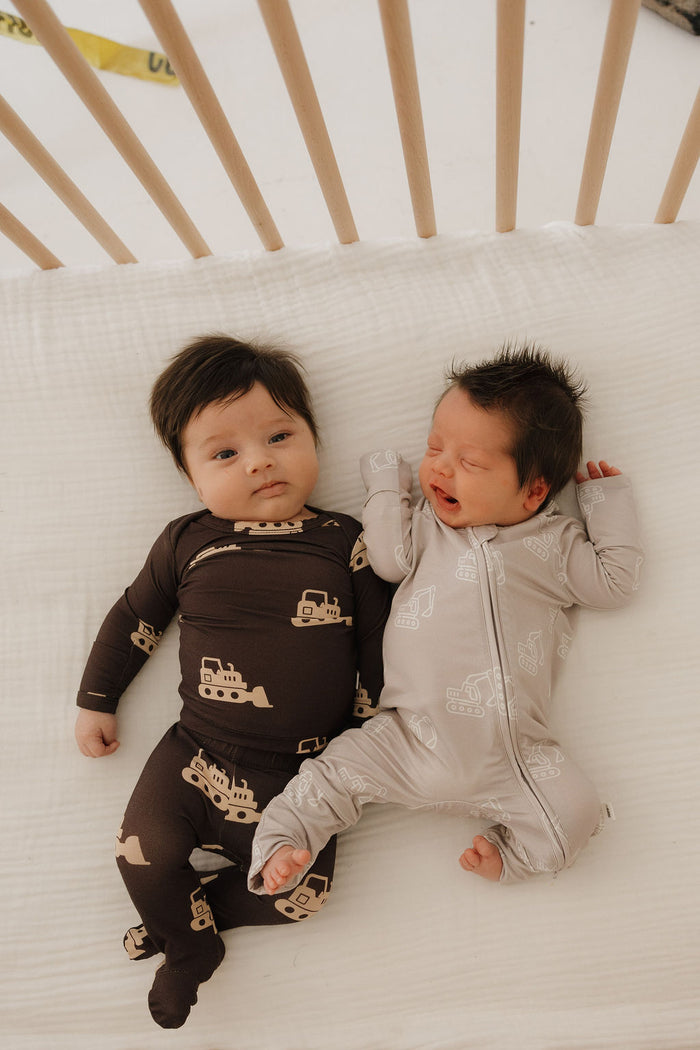 Two babies lie side by side in a crib. One, awake in forever french baby's Sleepy Time Set | Bulldozer pajamas, and the other, drowsy in light bamboo clothing—both wear breathable outfits designed for sensitive skin.