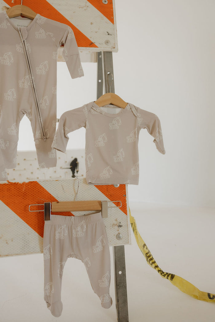 Three pieces from Forever French Baby’s Sleepy Time Set | Excavator, featuring hypoallergenic beige baby clothes with white construction vehicle prints, hang on wooden hangers before a striped orange-and-white metal sign and yellow caution tape.