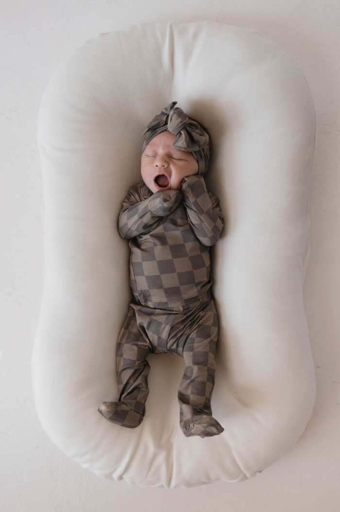 A baby wearing the Sleepy Time Set in Faded Brown Checkerboard from Forever French, along with a matching headwrap, lies on a white cushioned surface, yawning with arms raised towards the face. Clad in breathable, hypo-allergenic bamboo clothing, the baby is positioned on their back, seemingly relaxed and comfortable.