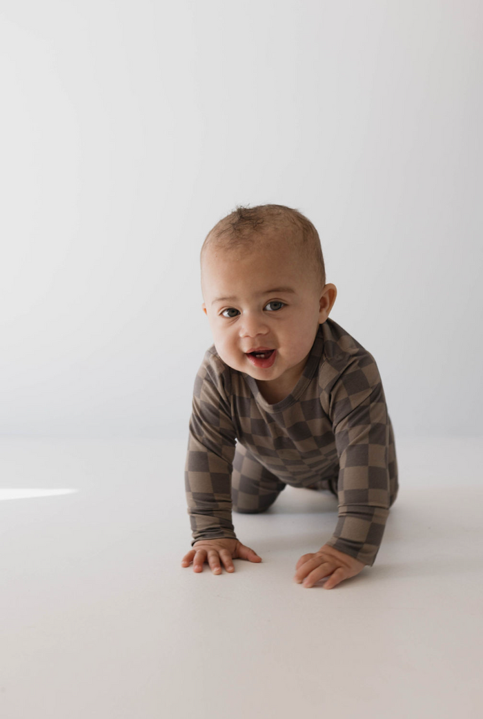 A baby wearing the hypo-allergenic, breathable "Sleepy Time Set | Faded Brown Checkerboard" onesie from Forever French is crawling on the floor against a white background, looking forward with a curious expression.