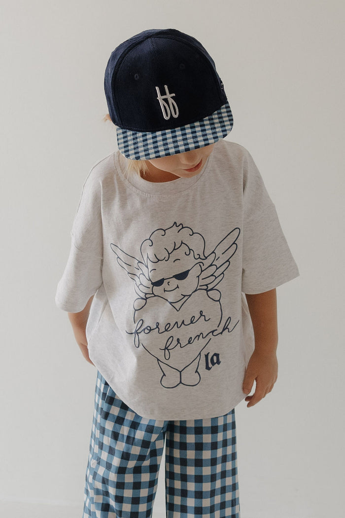 A child models the T-Shirt & Pant Set | ff Los Angeles by forever french baby, featuring a navy cap, loose light gray tee with a cartoon angel and "forever french" text, and blue gingham pants against a white background.