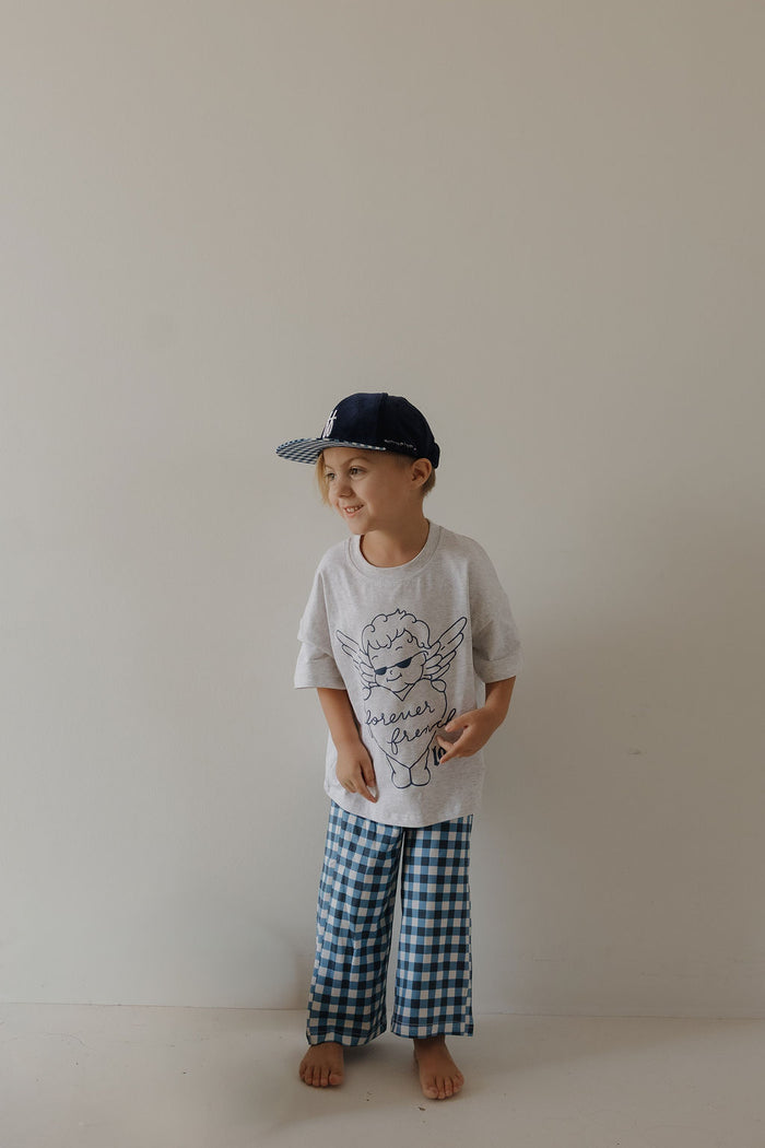 A young child wearing the forever french baby Snapback | Blue Corduroy ff stands barefoot against a plain light-colored wall, smiling and looking to the side.