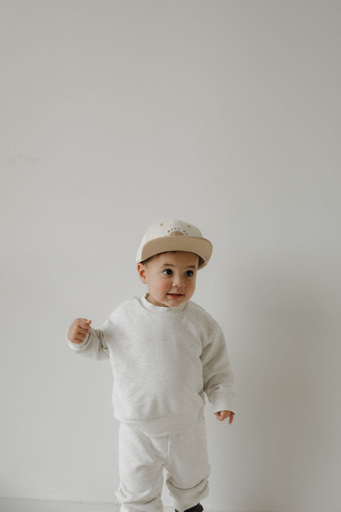 A toddler wears a "No Wake Zone" Snapback from forever french baby, standing against a plain white background and looking to the side with a curious expression.