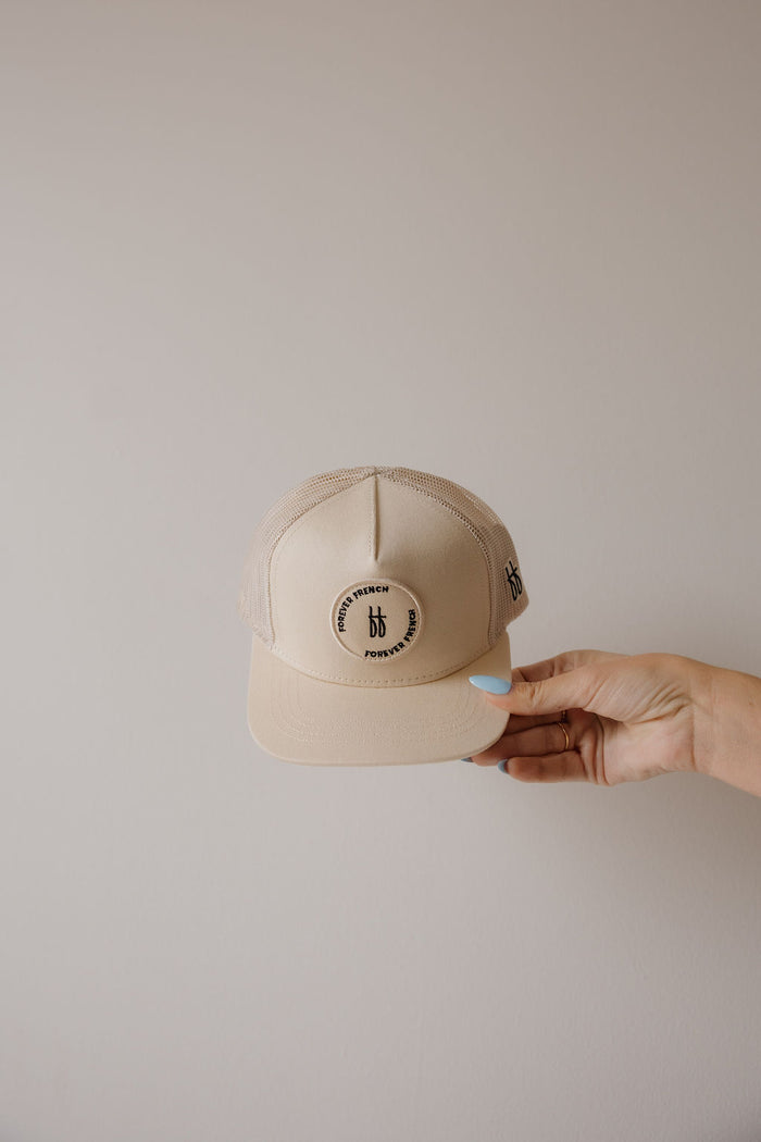 A hand with light blue polish holds the forever french baby Snapback | Sand, a beige trucker hat with a round logo patch displaying “Brun Blend,” “Portland, OR,” and a central “bb” monogram. The background is plain light gray.
