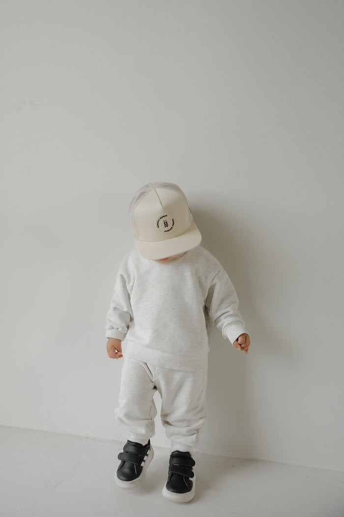 A small child in a light gray sweatshirt, sweatpants, black sneakers, and the Snapback | Sand from forever french baby stands against a white wall, looking down.