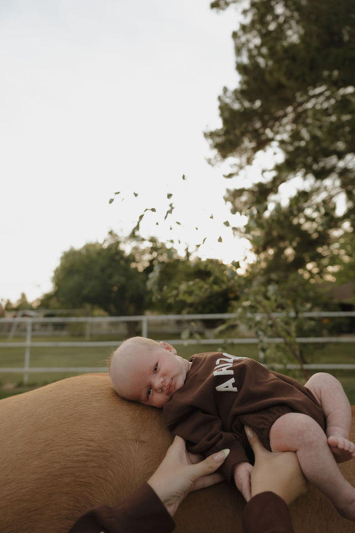 A baby in a forever french baby Sweatshirt Romper | ff Arizona is held by two hands, resting on a light brown animal outdoors with a fence and trees in the background.