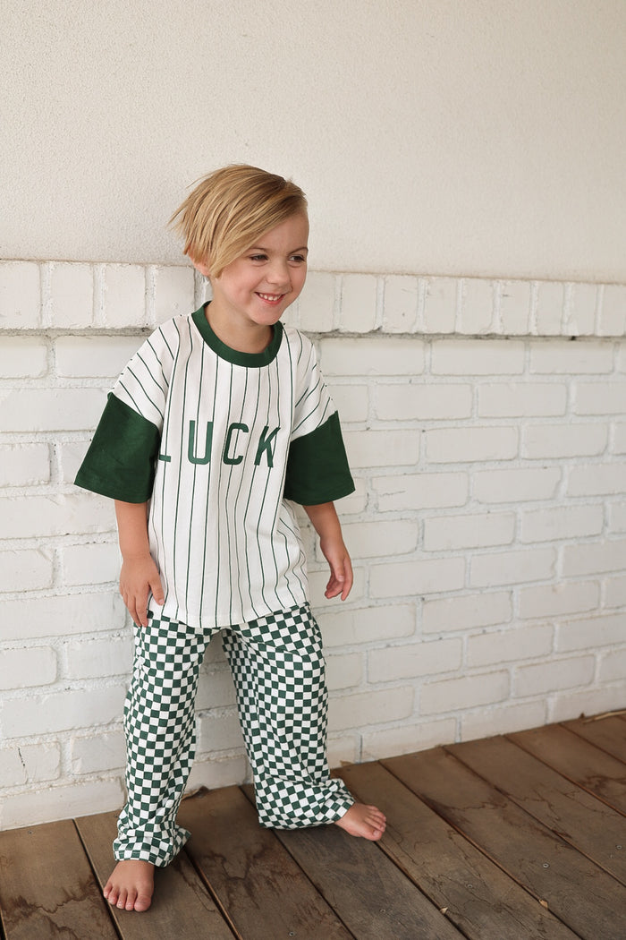 A smiling young child with blonde hair stands barefoot, wearing the forever french baby T-Shirt & Pant Set | Lucky Micro Check—featuring a white and green striped “LUCK” shirt and green wide leg pants—against a white brick wall.
