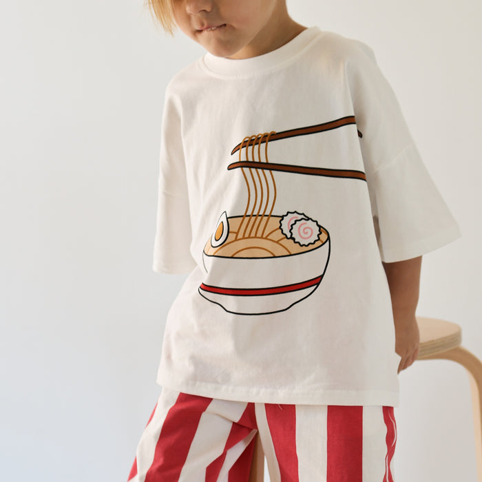 A child models the forever french baby T-Shirt & Pant Set | Ramen, featuring a cotton tee with a cartoon ramen bowl and chopsticks graphic, paired with red and white striped pants—a playful kids’ daywear set.
