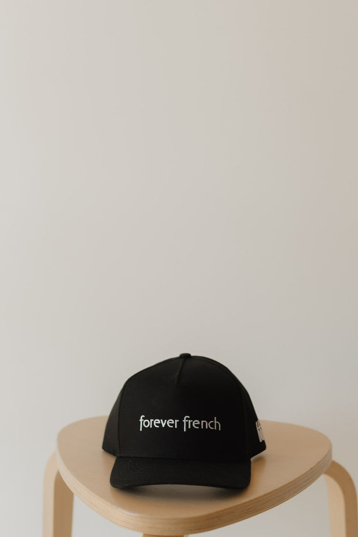 A Snapback | forever french Black by forever french baby, featuring white "forever french" logo stitching, rests on a light wooden stool against a plain, neutral background emphasizing its style.