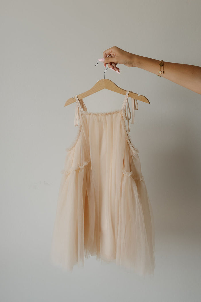 A hand holds a hanger with the forever french baby Tutu Dress in the Slipper shade, a light peach, sleeveless tulle tutu dress featuring delicate bow straps, set against a plain white background.