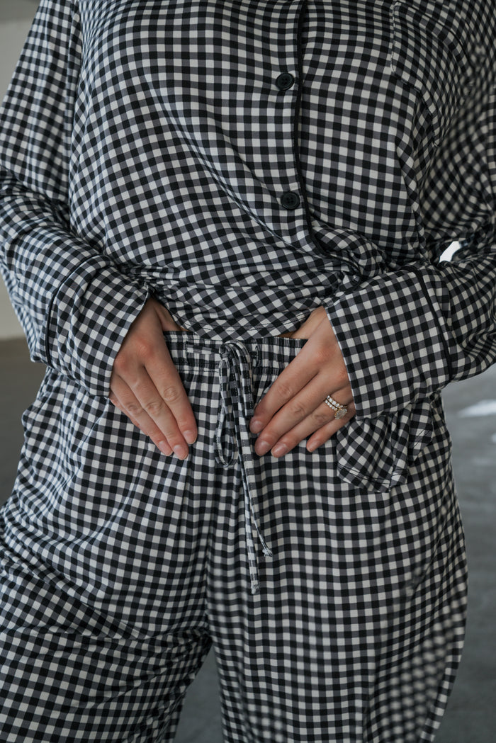 A person is shown wearing the Women’s Bamboo Button Up Lounge Set in Black Gingham by forever french baby, with hands resting on their stomach and rings adorning their left hand. The image focuses on the torso and lower part of the face, drawing attention to the soft bamboo material and relaxed fit of the wide-leg lounge pants.