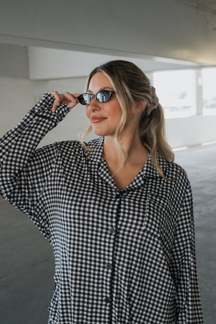 A woman in the forever french baby Women’s Bamboo Button Up Lounge Set in Black Gingham adjusts her sunglasses while standing in a bright, indoor parking area, effortlessly showcasing the comfort and relaxed style of bamboo clothing.