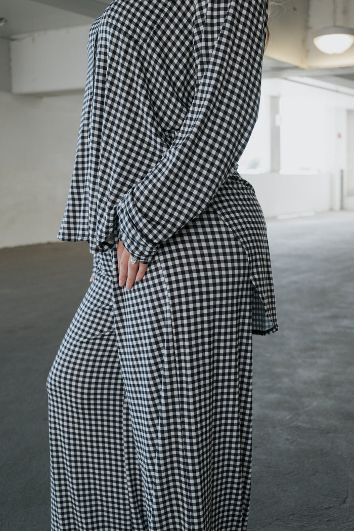 A person wearing the forever french baby Women’s Bamboo Button Up Lounge Set in Black Gingham stands indoors with one hand on their hip. The background features an empty, white-walled space illuminated by natural light.
