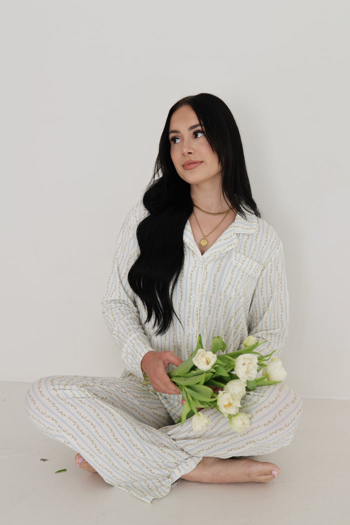 A woman with long dark hair sits cross-legged in the forever french baby Women's Bamboo Button Up Lounge Set | Dainty Vines, holding white tulips and gazing thoughtfully to the side against a plain white background.