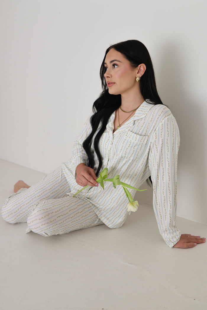 A woman with long dark hair sits on the floor in a forever french baby Women's Bamboo Button Up Lounge Set | Dainty Vines, holding a white tulip and enjoying the comfort of this hypo-allergenic bamboo loungewear.