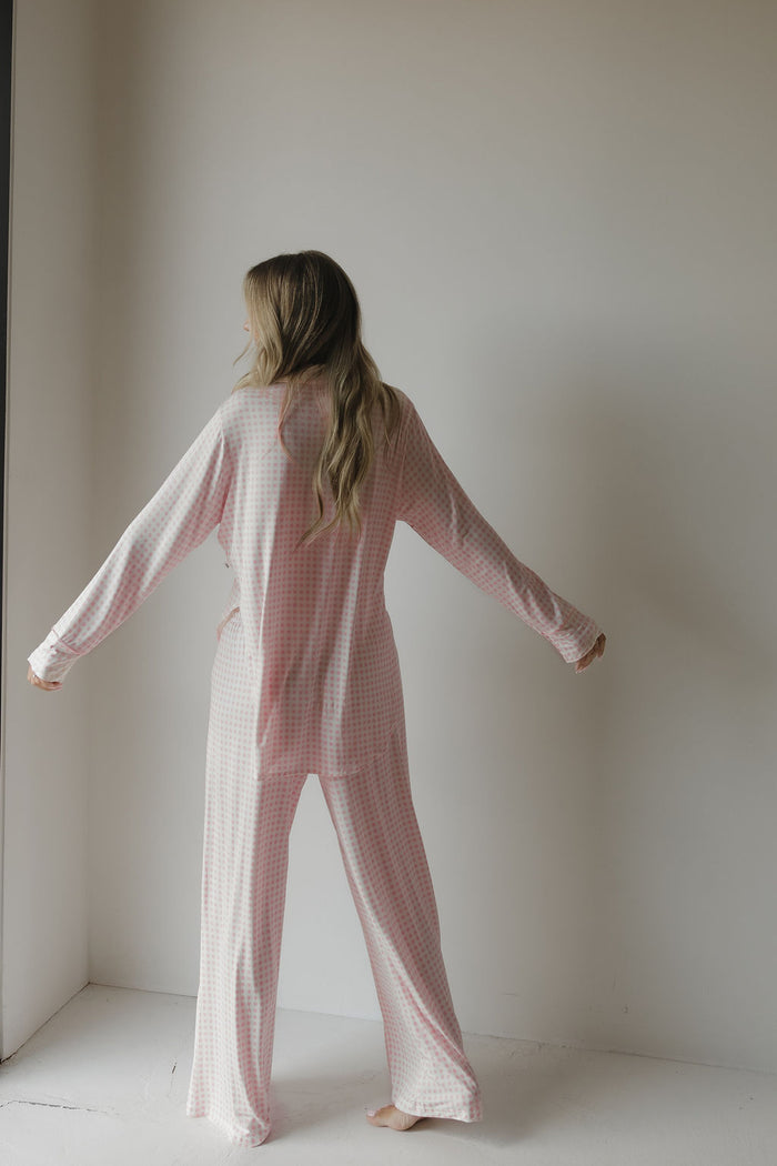 A woman with long hair stands barefoot facing a white wall, arms slightly outstretched, wearing the forever french baby Women’s Bamboo Button Up Lounge Set | Giselle in a loose pink and white checkered pattern.