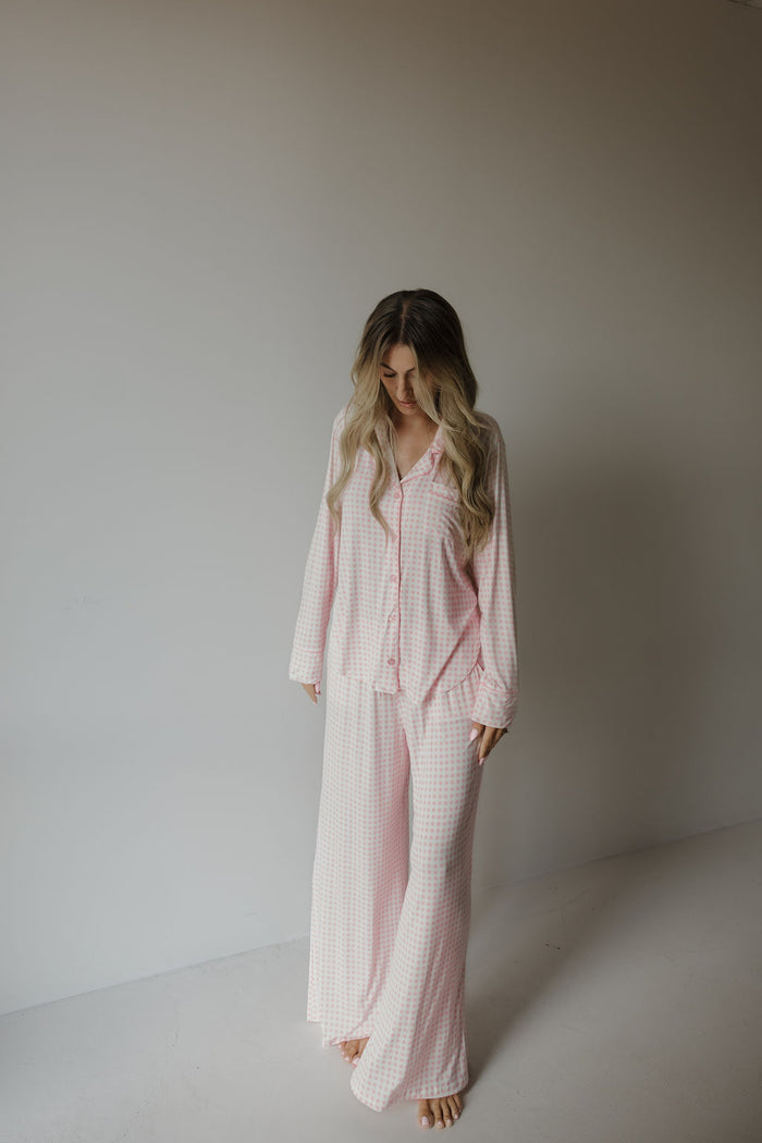 A woman with long, wavy hair stands barefoot indoors, wearing the forever french baby Women’s Bamboo Button Up Lounge Set | Giselle in a pink and white checkered pattern. She looks down as she touches her hair, enjoying the comfort and softness of this hypo-allergenic loungewear—perfect for relaxing at home.
