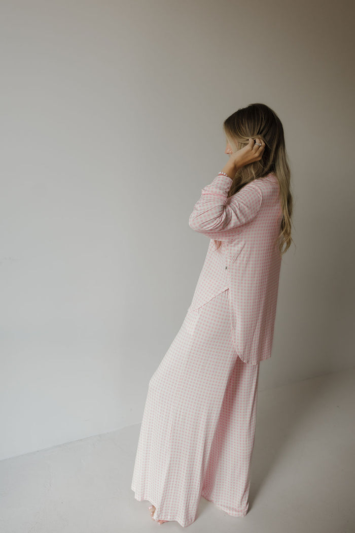 A person with long hair stands in a corner, wearing the Giselle Women’s Bamboo Button Up Lounge Set by forever french baby. They are dressed in a light pink and white checkered set from the women’s bamboo clothing collection, facing sideways and touching their hair against a neutral background.