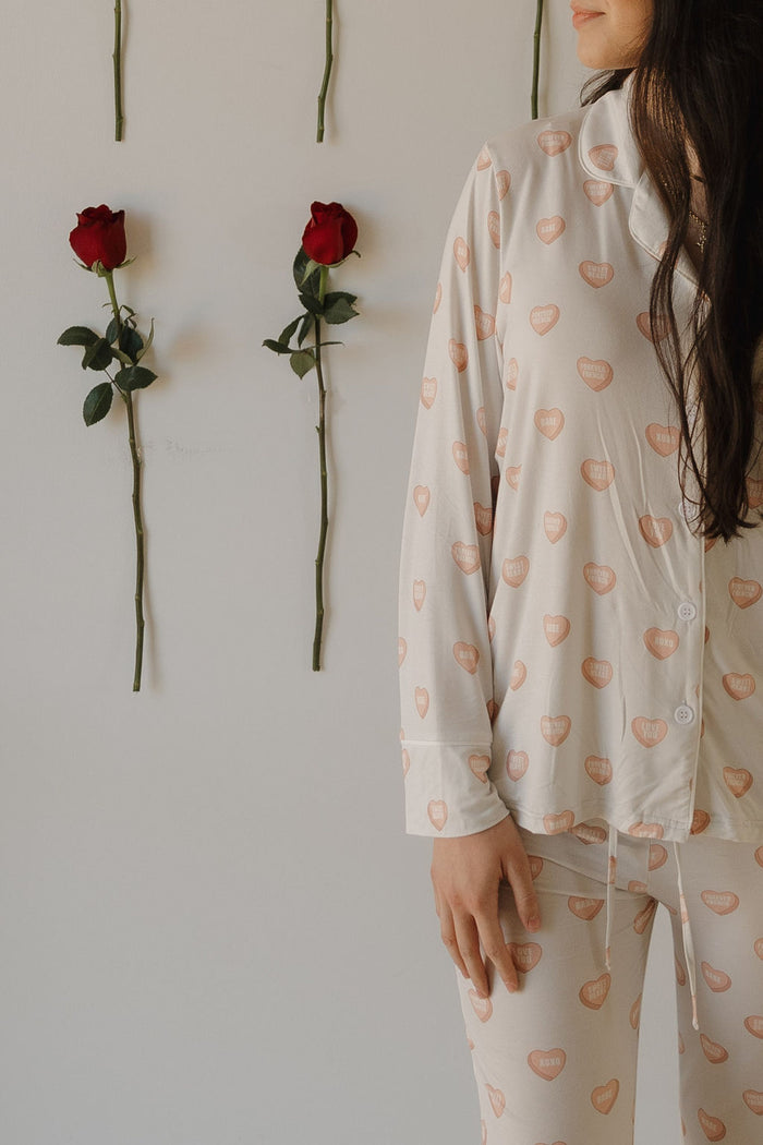 A woman, with only her lower face and torso visible, wears the forever french baby Women's Bamboo Button Up Lounge Set | Love Notes - Grey as she stands by a white wall decorated with three red roses.