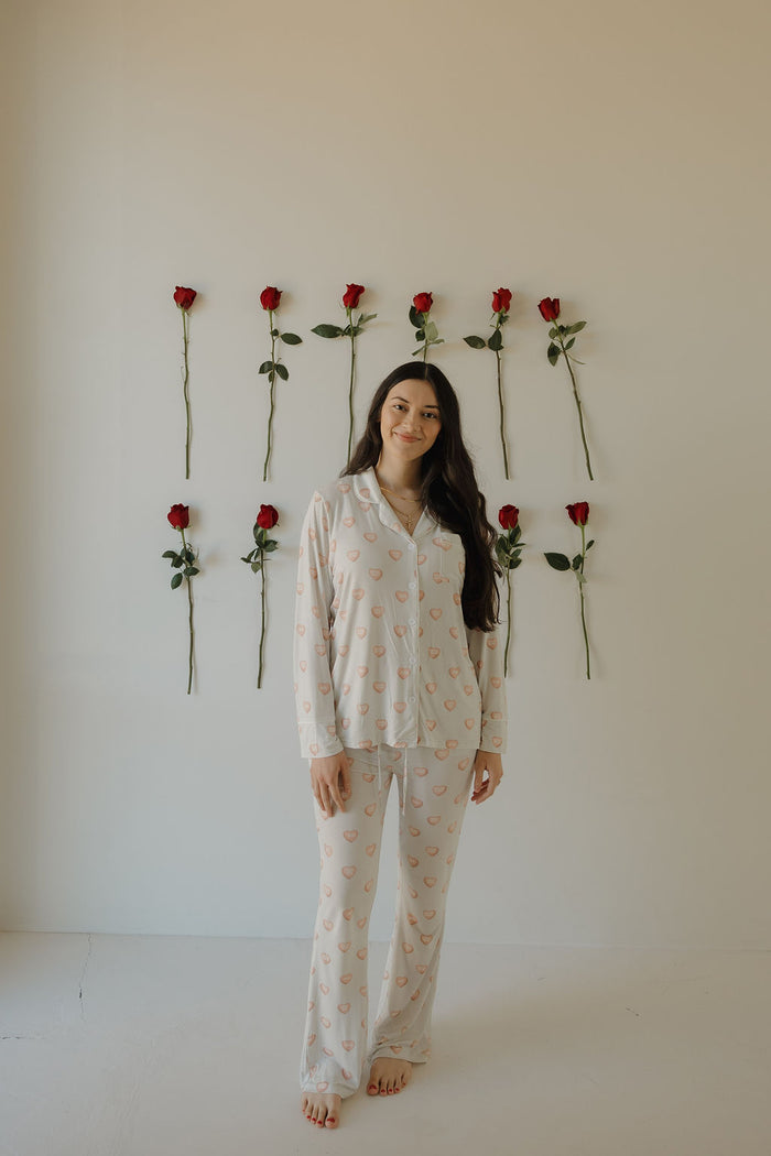 A woman with long dark hair stands barefoot, smiling in front of a white wall with eight vertically arranged red roses, wearing forever french baby's Women's Bamboo Button Up Lounge Set | Love Notes - Grey.
