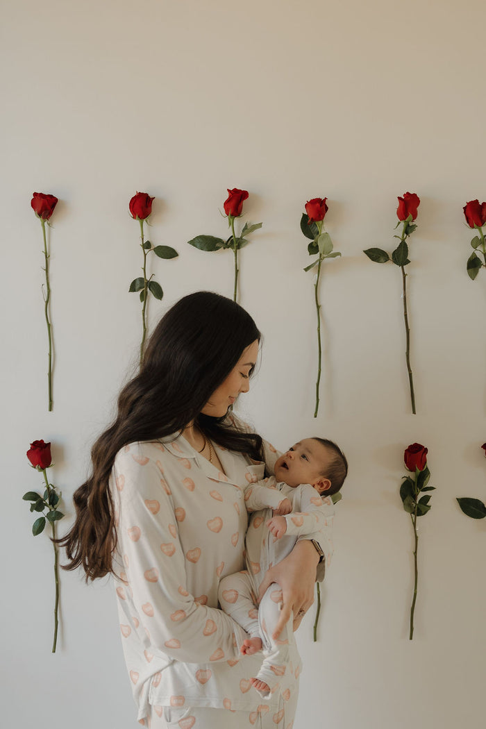 A woman wears the forever french baby Women's Bamboo Button Up Lounge Set | Love Notes - Grey while holding a baby, smiling in front of a white wall decorated with rows of red roses, highlighting stylish, hypo-allergenic bamboo clothing.