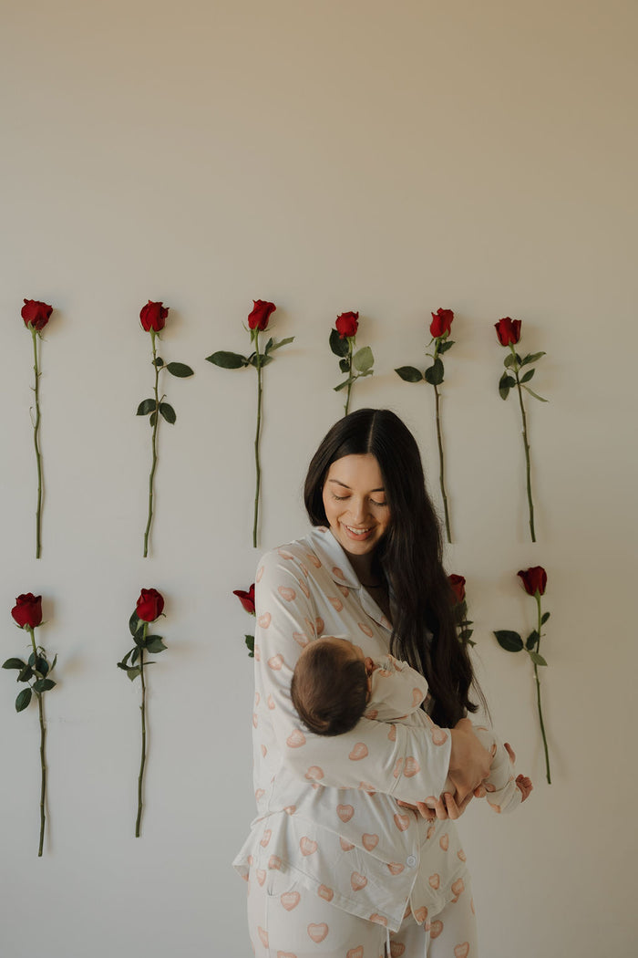 A smiling woman in the forever french baby Women's Bamboo Button Up Lounge Set | Love Notes - Grey gently holds a baby. Red roses arranged in three rows decorate the plain wall, creating a warm, serene scene that highlights cozy bamboo loungewear.