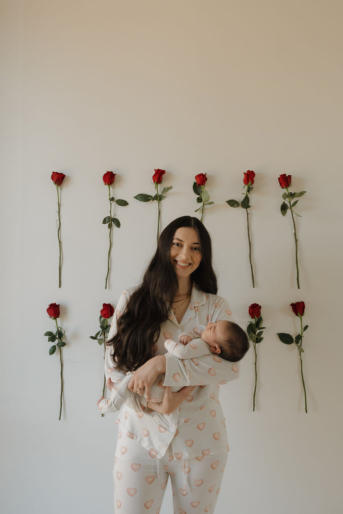 A woman in the forever french baby Women's Bamboo Button Up Lounge Set | Love Notes - Grey holds a baby, both looking content and relaxed in soft, hypo-allergenic bamboo clothing, standing before a white wall with rows of red roses.