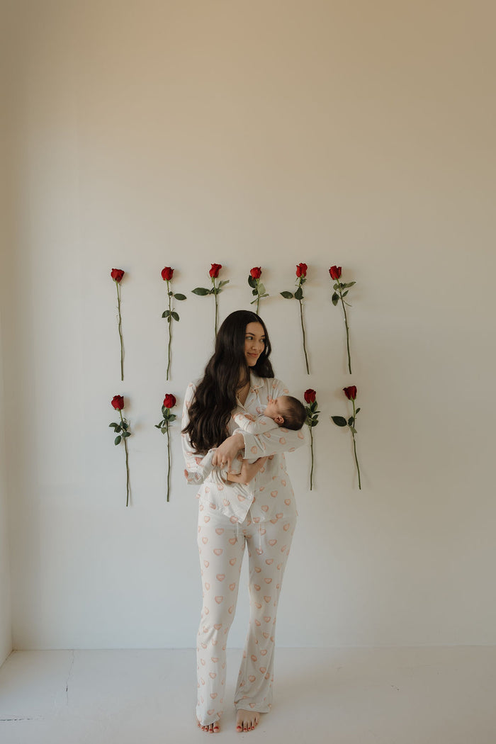 A woman in the forever french baby Women's Bamboo Button Up Lounge Set | Love Notes - Grey holds a baby, both barefoot by a white wall with nine red roses—enjoy everyday comfort and style in hypoallergenic bamboo clothing.