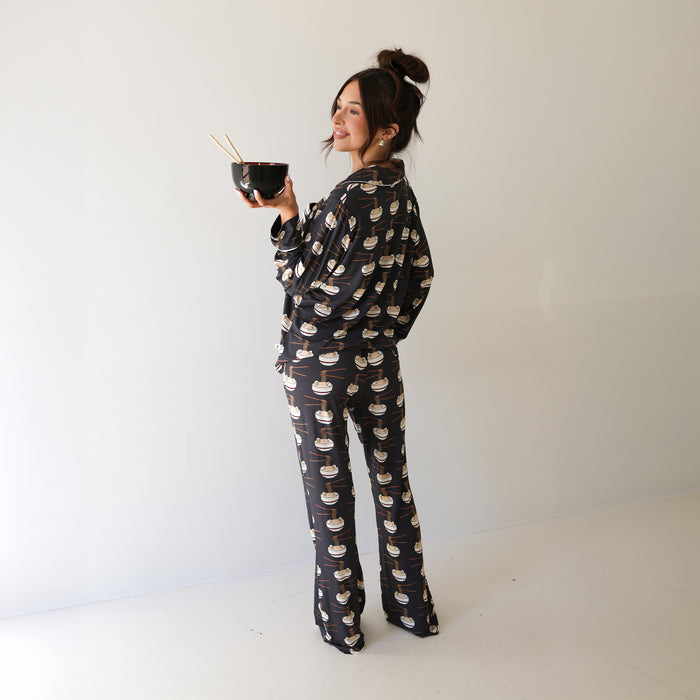 A woman with a bun smiles sideways, holding a bowl with chopsticks and wearing the forever french baby Women's Bamboo Button Up Lounge Set | Ramen—hypo-allergenic bamboo clothing with a fun ramen bowl pattern against a light-colored wall.