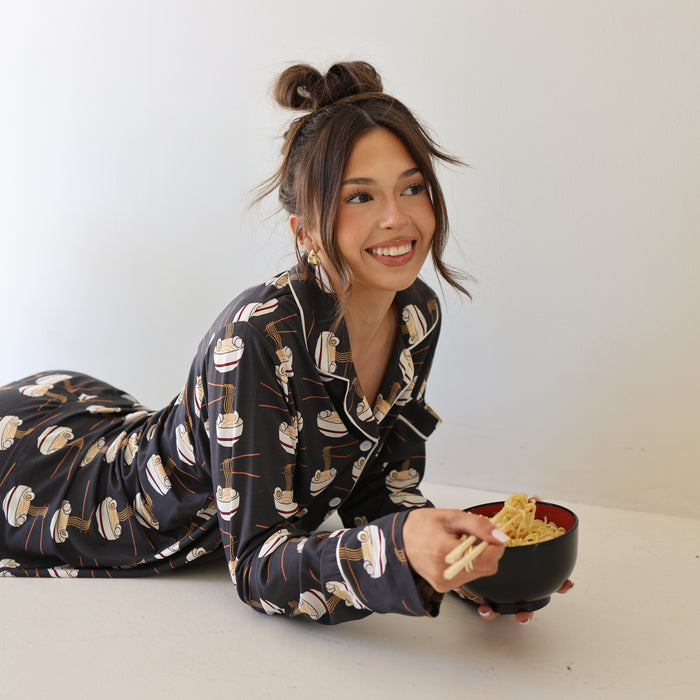 A woman smiles and lounges on her side in the forever french baby Women's Bamboo Button Up Lounge Set | Ramen, featuring patterned ramen bowls. She holds a bowl of noodles with chopsticks against a plain, light background.