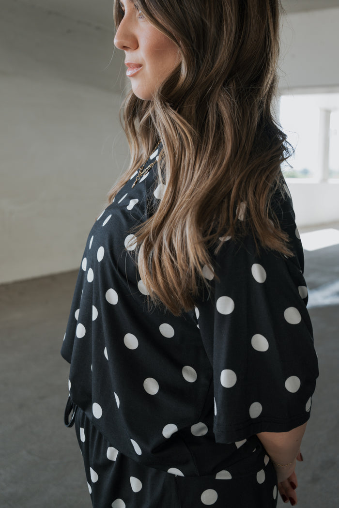 A woman with long brown hair stands in a bright room, facing sideways with her hands behind her back, wearing the forever french baby Women’s Bamboo Short Set in black and white polka dot—made from breathable bamboo fabric.
