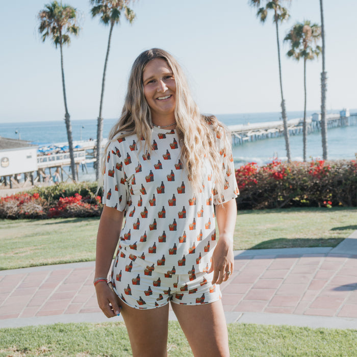 A woman with long, wavy hair smiles outdoors by the ocean, wearing forever french baby's Women's Bamboo Short Set in the Fries & Cola print. Palm trees and a pier are visible in the sunny background.