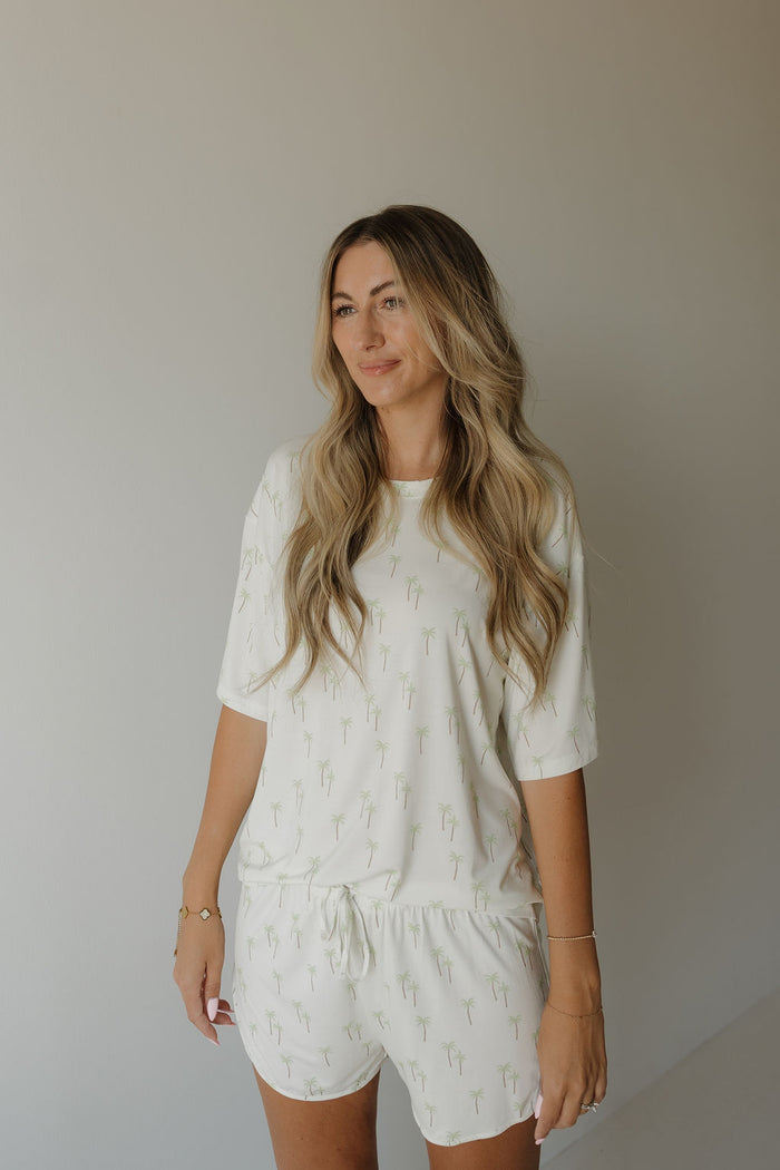 A woman with long wavy blonde hair wears the forever french baby Women’s Bamboo Short Set | Palm—a breathable bamboo pajama set featuring subtle small green palm tree prints—as she stands against a plain light-colored wall and smiles softly.
