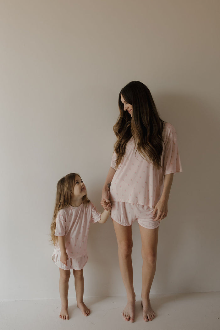 A woman and a young girl, both with long brown hair, stand barefoot against a plain wall, holding hands and smiling at each other while wearing matching forever french baby Women’s Bamboo Short Set | Rosie pajamas in light pink polka dots, made from soft, breathable bamboo fabric.