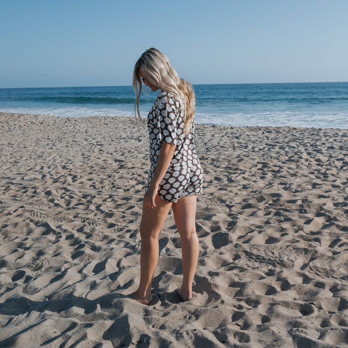 A woman with long blonde hair wears the Women's Bamboo Short Set | Seashells by forever french baby, standing barefoot on a sandy beach with the ocean and blue sky in the background.