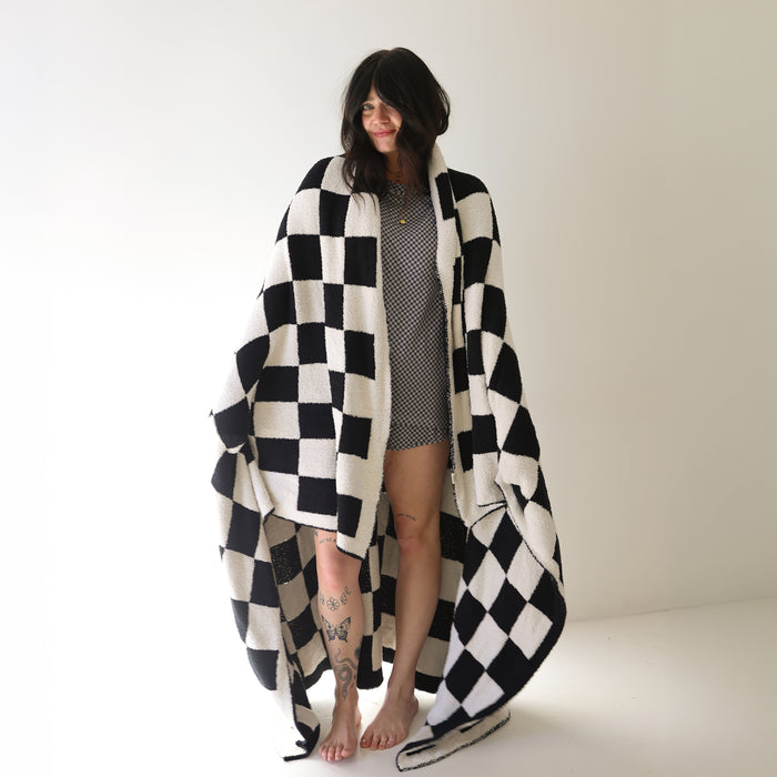 A woman, barefoot in a bright room, is wrapped in the forever french baby Plush Blanket | Black & White Checkerboard. She wears a short patterned dress, has tattoos on her legs, and smiles gently while looking to the side.