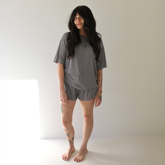 A woman with long dark hair and visible tattoos stands barefoot in a well-lit room, smiling slightly while wearing the forever french baby Women's Bamboo Short Set | Street Micro Check.
