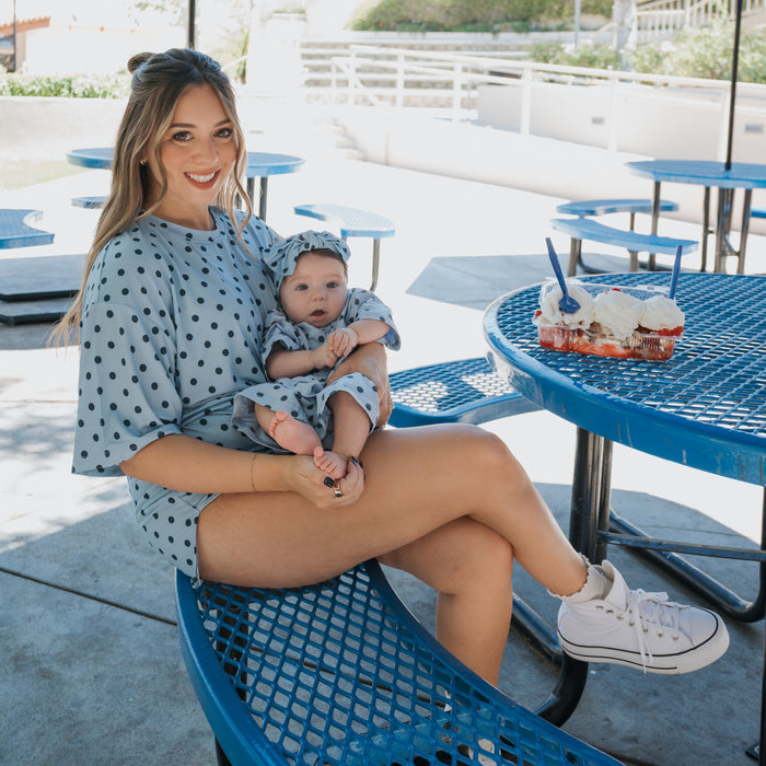 A woman wears the forever french baby Women's Bamboo Short Set | Sully in a blue polka-dot print while holding a matching baby, both seated outdoors at a blue picnic table with three frosted cupcakes and two spoons.