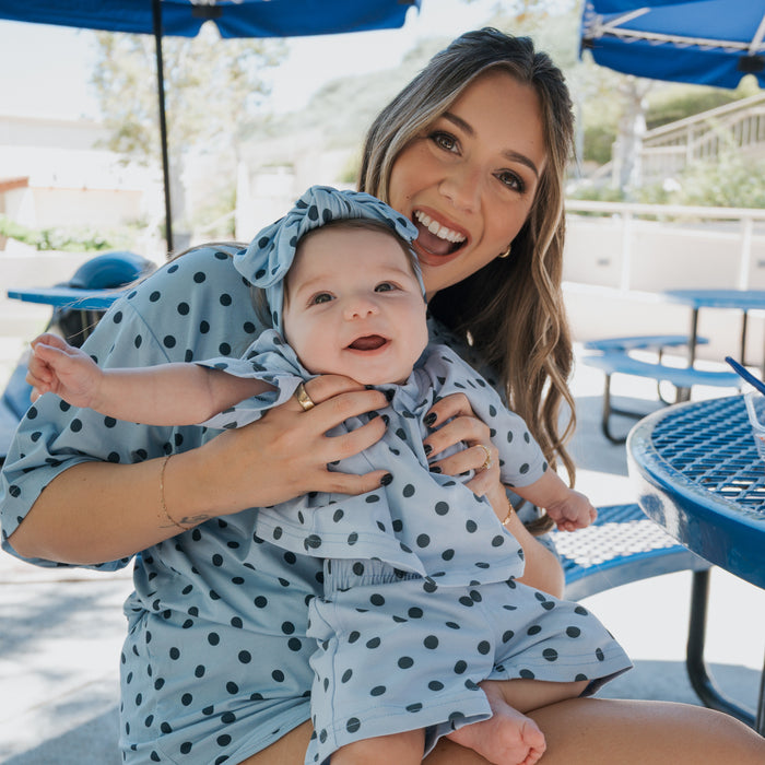 A smiling woman wearing the forever french baby Women's Bamboo Short Set | Sully in blue polka dots holds a happy baby dressed in matching hypo-allergenic bamboo clothing, as they sit together at a blue outdoor table on a sunny day.