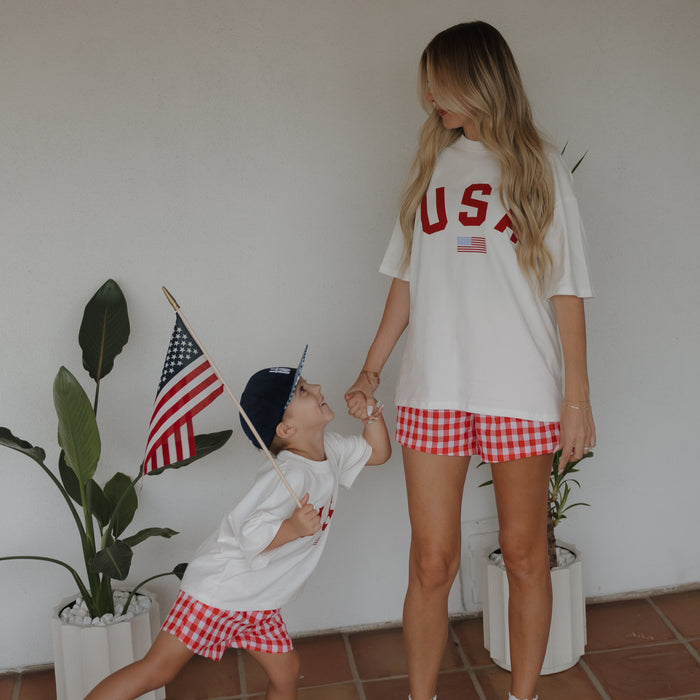 A woman and child, both in matching "USA" shirts and forever french baby's Women's Boxer Shorts | Scarlet Gingham, hold hands indoors by potted plants. The child, holding an American flag, looks up at the woman.