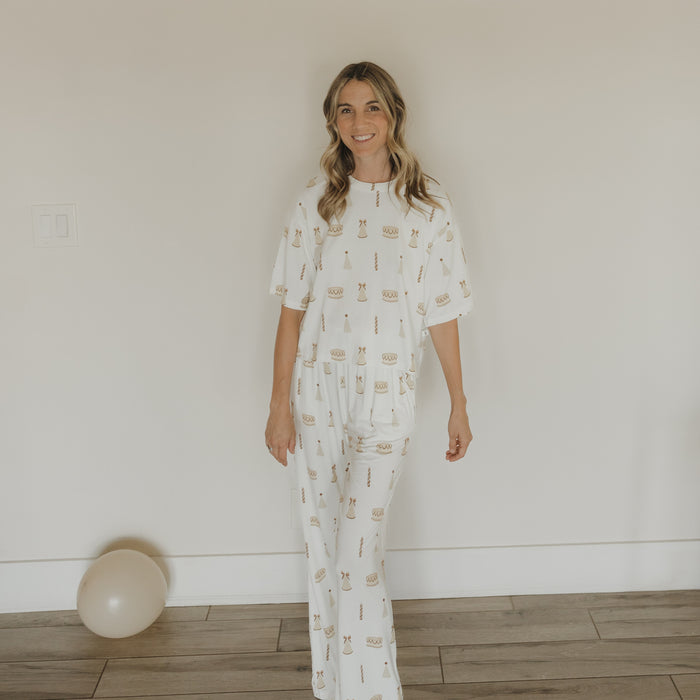 A woman, smiling, stands indoors on a wooden floor near a white wall. She is wearing the "Women's Boxy Tee Lounge Set | Birthday Girl" by forever french baby—a cozy and stylish choice for relaxing at home. In the corner, a single white balloon rests on the floor.
