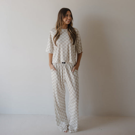A woman with long, wavy hair stands against a white wall wearing the forever french baby Women's Boxy Tee Lounge Set | Checkerboard, featuring a beige and white checkered pattern, short sleeves, and loose-fitting pants with her hands in her pockets.