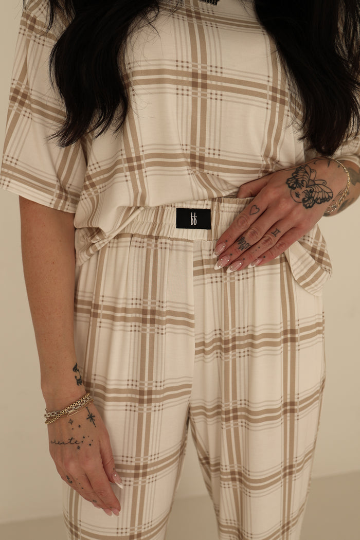 A woman models the Women's Boxy Tee Lounge Set in Ellis Plaid by forever french baby, showing tattoos and a gold bracelet. Only her torso and hands are visible. The hypo-allergenic outfit features a black "bb" tag for style and comfort.