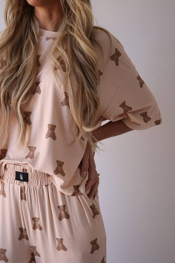 A person wears the forever french baby Women's Boxy Tee Lounge Set | Lottie, a matching beige set with a brown teddy bear print. The loose short-sleeve top and elastic waist pants complete the cozy look, with long wavy hair draped over one shoulder.