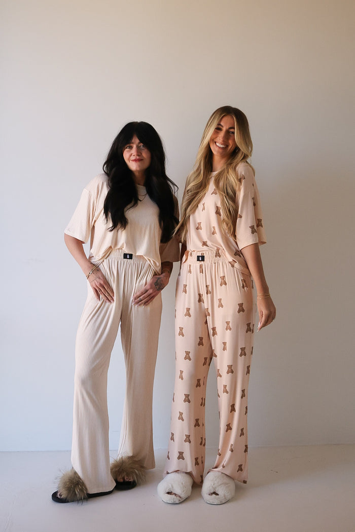 Two women smile side by side in matching forever french baby Women's Boxy Tee Lounge Set | Lottie—one in cream, the other in pink with a brown teddy bear print—paired with fluffy slippers, against a plain light background.